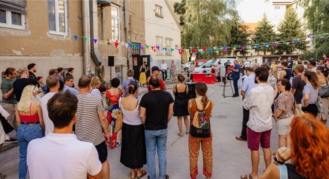 Slika prikazuje skupino ljudi, ki se udeležujejo dogodka na prostem, kjer so razvrščeni okoli govorniškega odra.