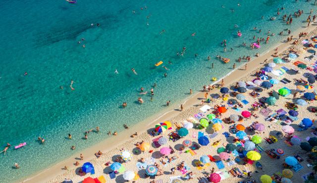 Pogled od zgoraj na živahno plažo z množico pisanih senčnikov in kopalcev v turkiznem morju.